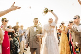 A beautiful wedding photo taken at Bodrum ruins featuring a happy bride and groom exiting the outdoor ceremony amidst a shower of confetti and flower petals. The couple stands side by side, smiling, and surrounded by their guests. 